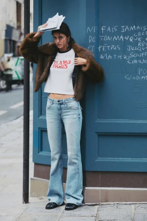 Woman in brown fur coat and jeans leaning against blue door, holding papers overhead, in urban setting. Fashion editorial style.