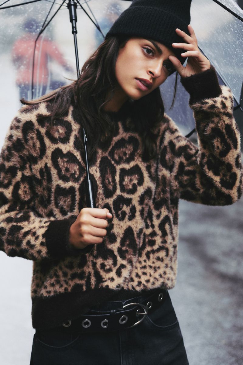 Brunette woman in an animal-print sweater and black beanie stands beneath a clear umbrella on a city street on an overcast day.