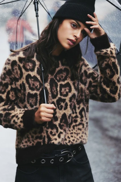 Brunette woman in an animal-print sweater and black beanie stands beneath a clear umbrella on a city street on an overcast day.