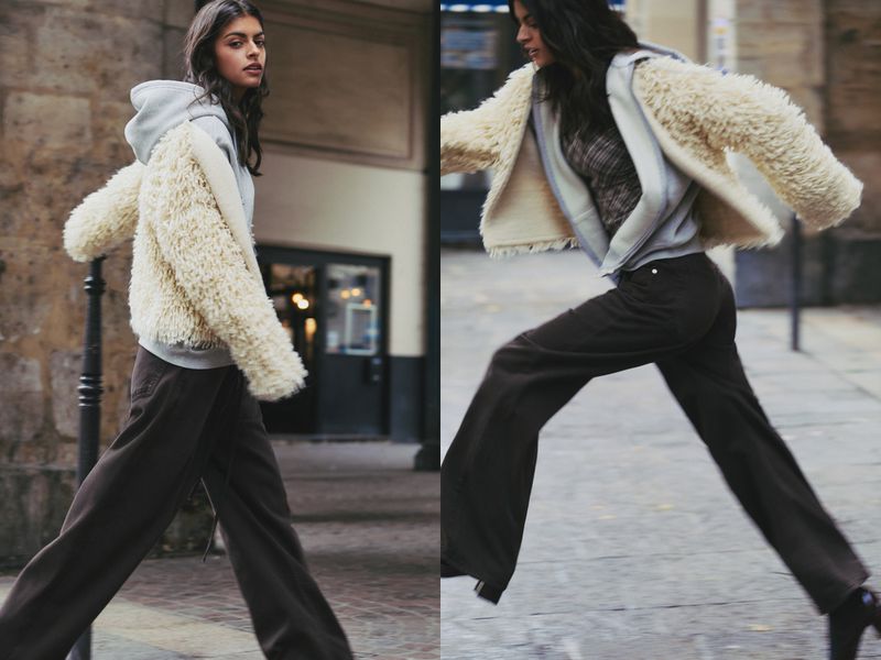 A brunette woman in motion on a cobblestone street wearing brown pants, a light hooded sweatshirt, and a fuzzy short cream-colored jacket.