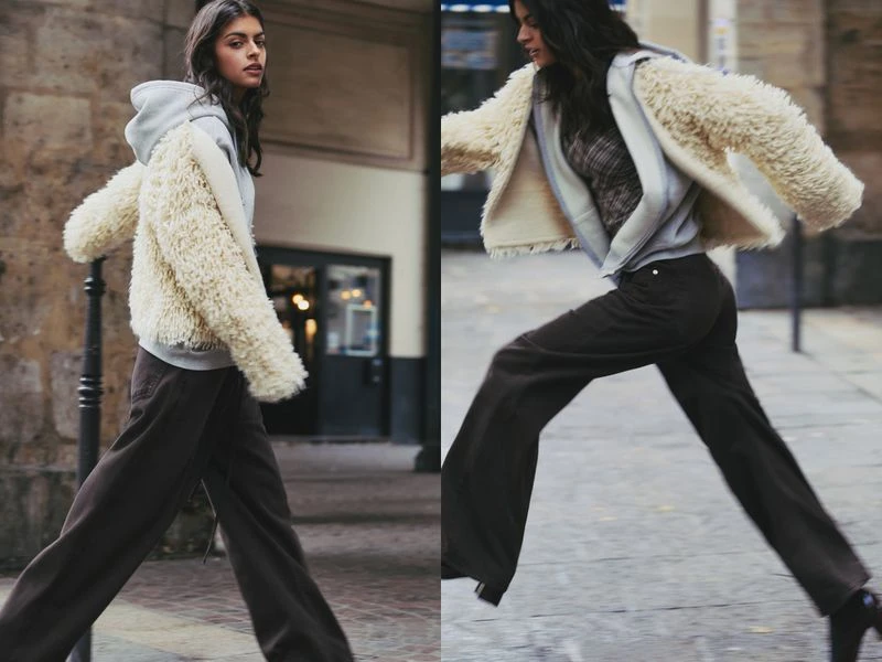 A brunette woman in motion on a cobblestone street wearing brown pants, a light hooded sweatshirt, and a fuzzy short cream-colored jacket.