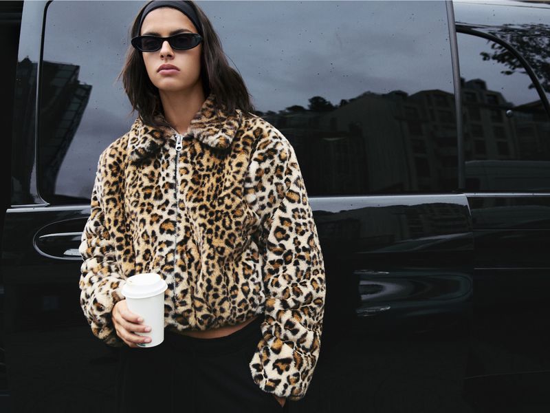A young woman wearing a leopard-print jacket and sunglasses stands in front of a glossy black vehicle, holding a paper cup.
