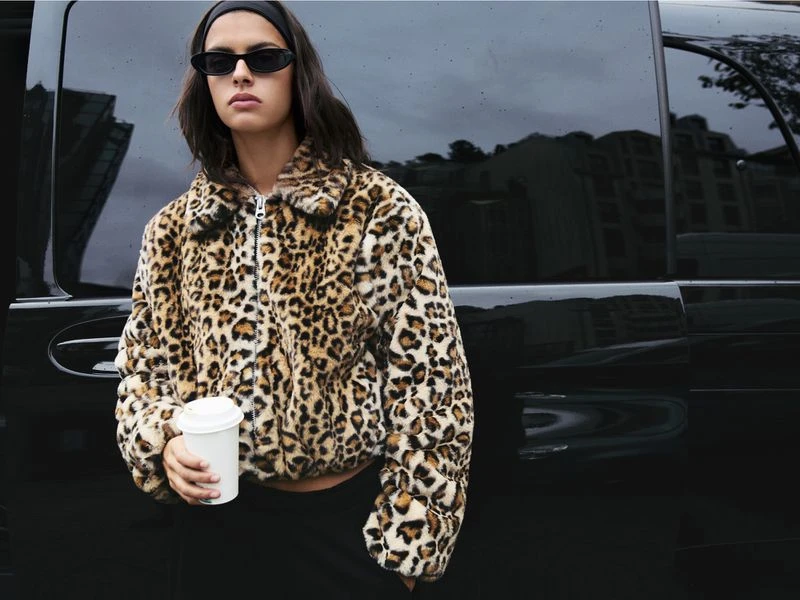 A young woman wearing a leopard-print jacket and sunglasses stands in front of a glossy black vehicle, holding a paper cup.