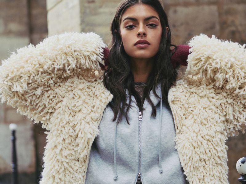 A young woman stands outdoors wearing a light-gray zip-up hoodie and a fluffy, ivory-colored shag coat with a stone wall background.