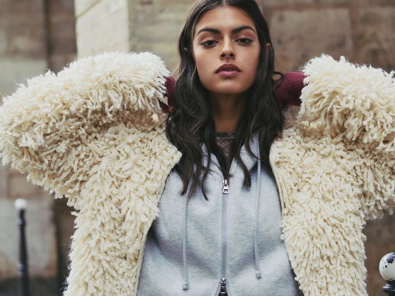 A young woman stands outdoors wearing a light-gray zip-up hoodie and a fluffy, ivory-colored shag coat with a stone wall background.