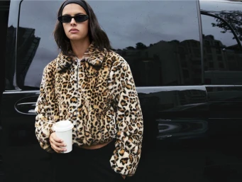 Model wearing a leopard print faux fur jacket, sunglasses, and headband, holding a coffee cup in front of a black car.