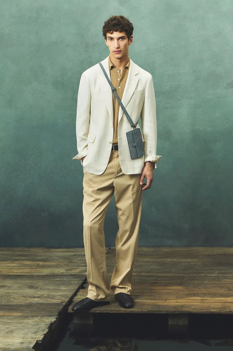 Model wearing an off-white blazer, tan polo shirt, and beige trousers posing on a wooden platform before a textured green backdrop.