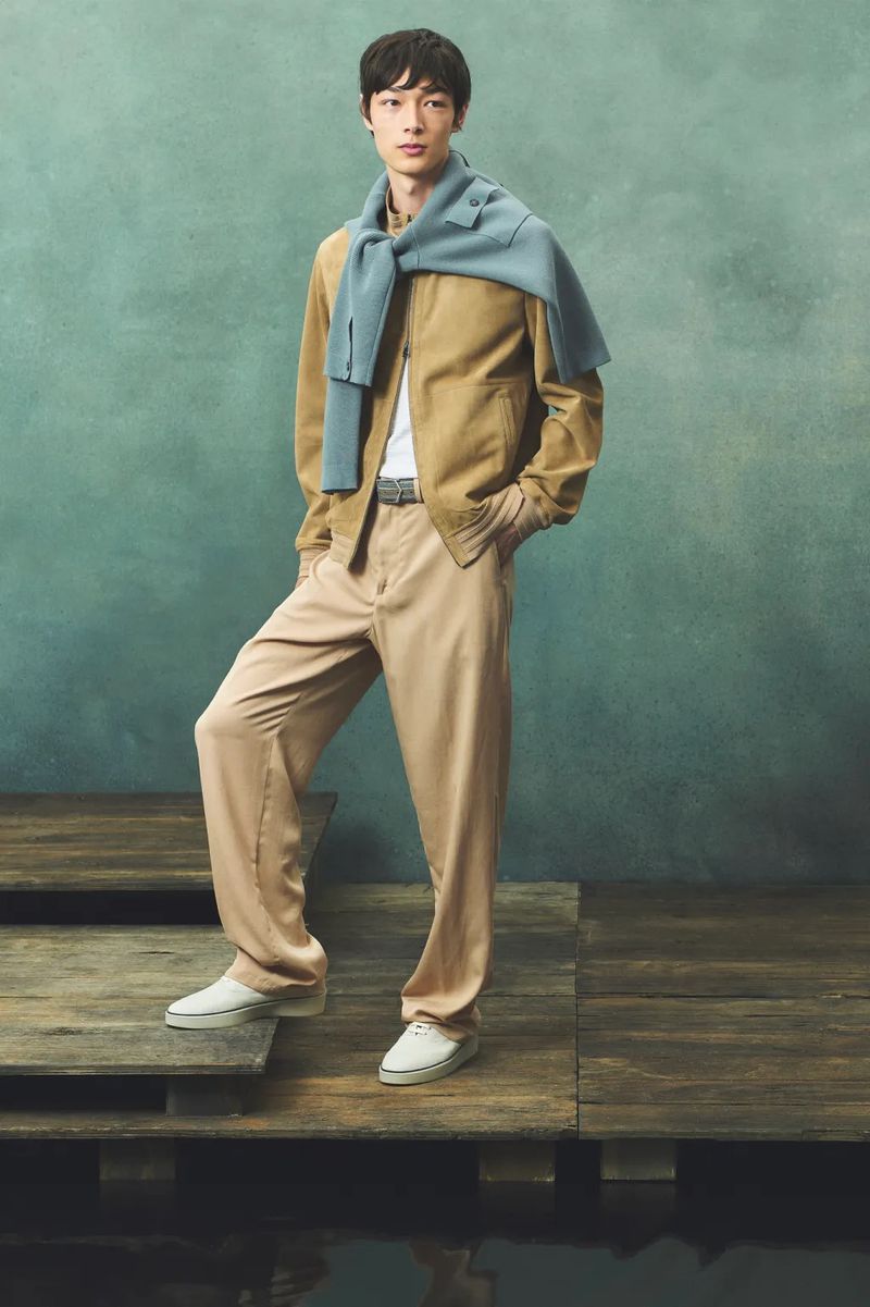 A stylish Asian male model poses in beige jacket and pants with a blue sweater draped over his shoulder, standing on a wooden platform.