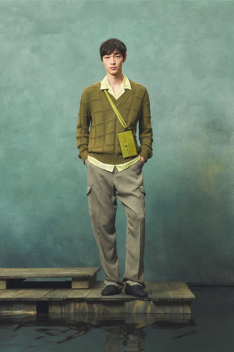 Full-shot studio portrait of a stylish Asian man in a sweater and cargo pants, standing on a wooden platform against a teal background.