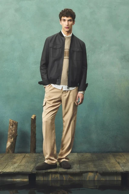 A man in a jacket, sweater, and khaki pants stands on a wooden platform near water, against a blue-green mottled background.
