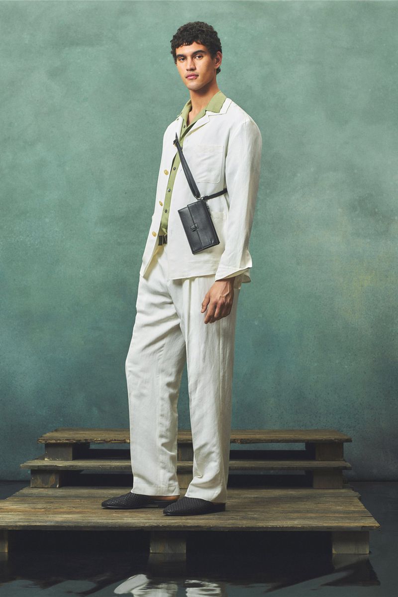 A male fashion model poses on a wooden platform wearing a white linen suit and black crossbody bag against a green textured backdrop.