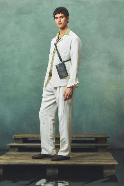 A male fashion model poses on a wooden platform wearing a white linen suit and black crossbody bag against a green textured backdrop.