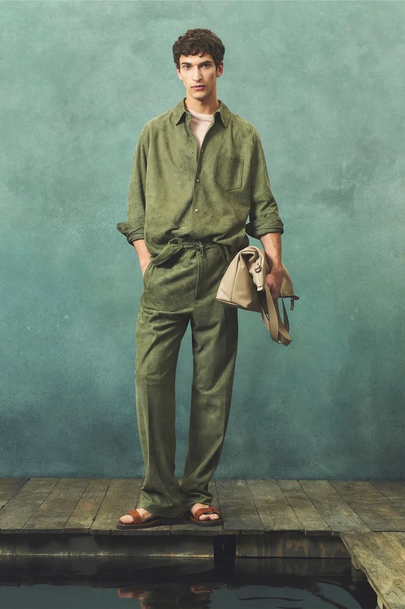 Male model poses on a wooden dock wearing an olive green linen suit, matching shirt and sandals, holding a canvas bag.