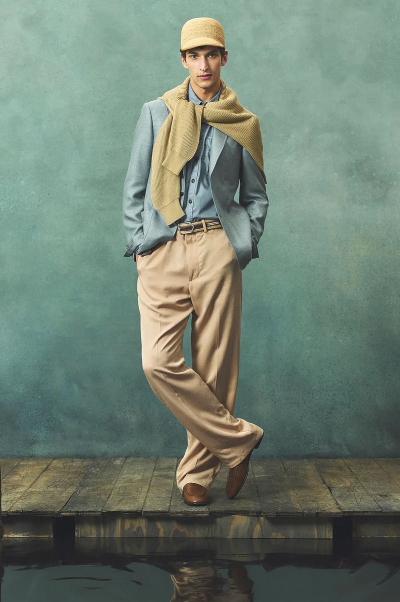 Male model wearing a blue suit, light brown pants, and a cap stands on a wooden dock against a blue backdrop with water reflections.