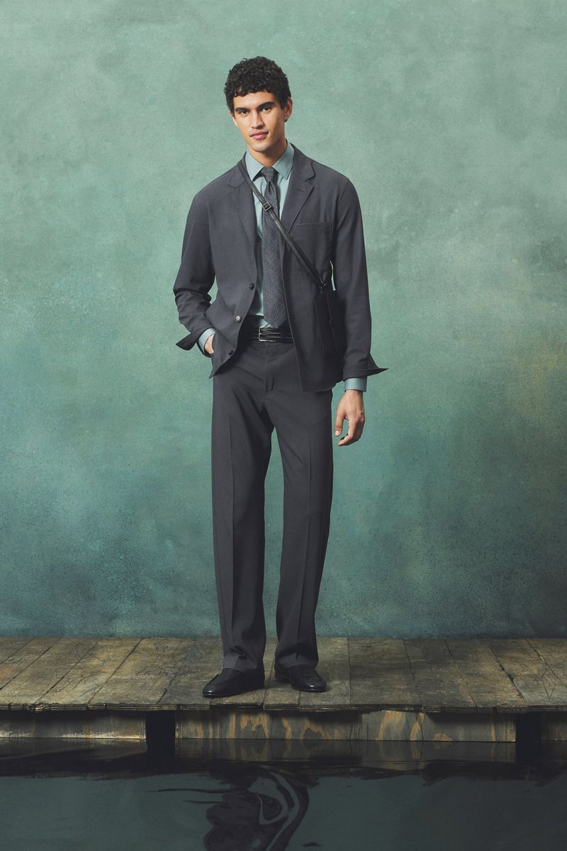 Fashionable adult male poses in a grey suit, light blue shirt, and tie on a wooden pier with water and a green mottled background.