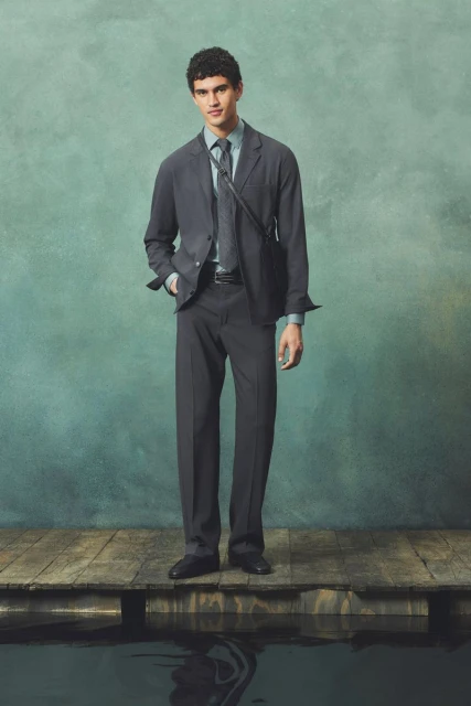 Fashionable adult male poses in a grey suit, light blue shirt, and tie on a wooden pier with water and a green mottled background.
