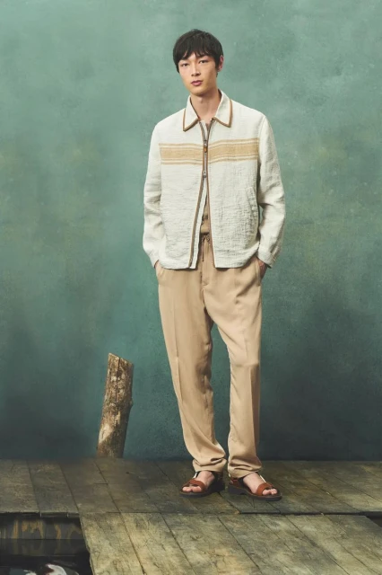 A stylish Asian man models fashion, wearing a cream jacket, tan pants, and sandals, posing on a wooden surface against a teal background.