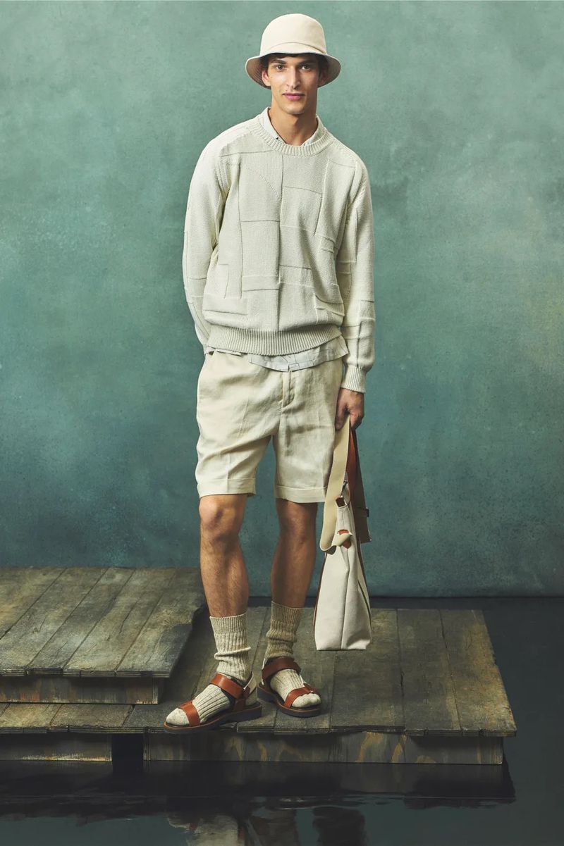 A slender man stands on a weathered wooden platform in front of a muted teal backdrop, modeling beige apparel and holding a tote bag.