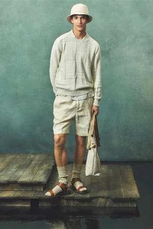 A slender man stands on a weathered wooden platform in front of a muted teal backdrop, modeling beige apparel and holding a tote bag.