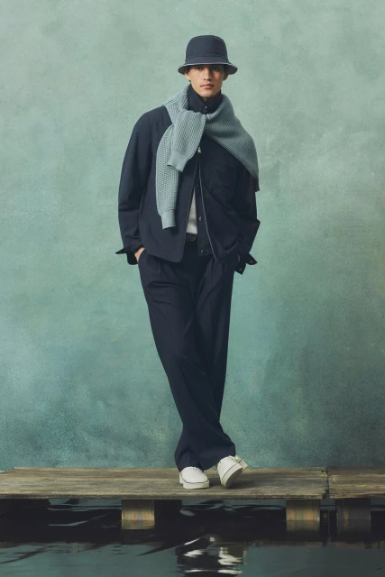 A full-length studio shot of a male model wearing a navy jacket, trousers, bucket hat, sweater, and white sneakers. He stands on a wooden dock.