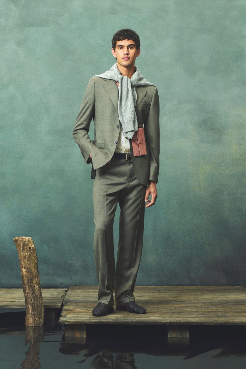 An adult male poses on a wooden dock wearing a grey suit, white shirt, and dark shoes against a muted teal background.