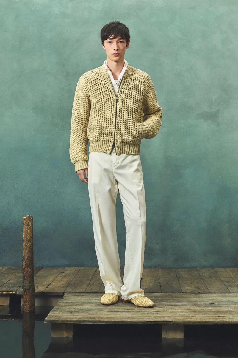 Full shot of a young adult man posing on a wooden pier in a textured tan sweater, white shirt, and white slacks against a blue-green backdrop.