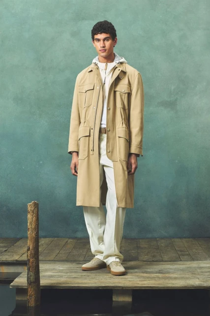 A man in a tan trench coat stands on a wooden dock with a muted green background, showcasing neutral-toned fashion in an outdoor setting.
