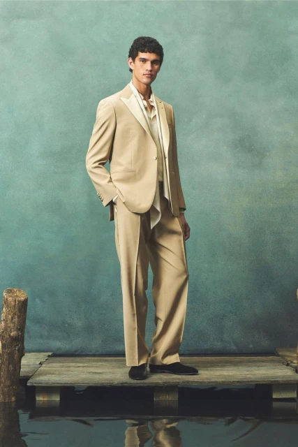 Fashionable adult man posing in tan suit with silk scarf, standing on rustic wooden dock, water and textured teal backdrop.