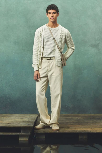 A young adult male poses in a light-colored knit sweater, cardigan, and pants on a wooden platform with a diffused green backdrop.
