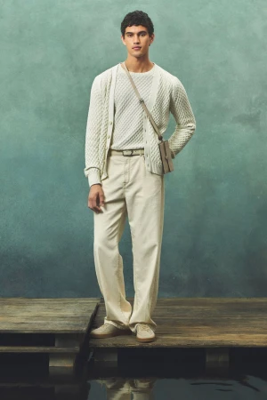 A young adult male poses in a light-colored knit sweater, cardigan, and pants on a wooden platform with a diffused green backdrop.