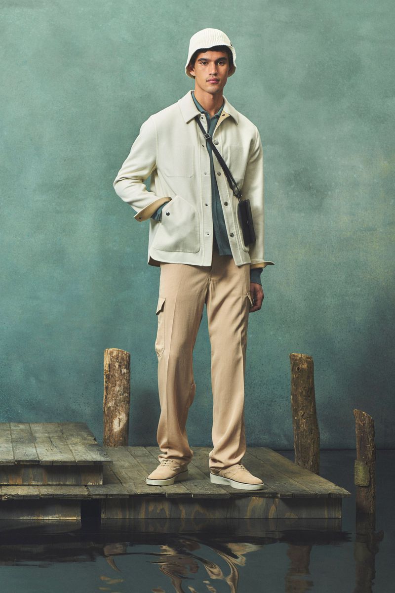 Male fashion model in beige coat, pants, bucket hat standing on a wooden dock against a mottled teal background, shoulder bag.