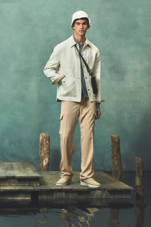 Male fashion model in beige coat, pants, bucket hat standing on a wooden dock against a mottled teal background, shoulder bag.
