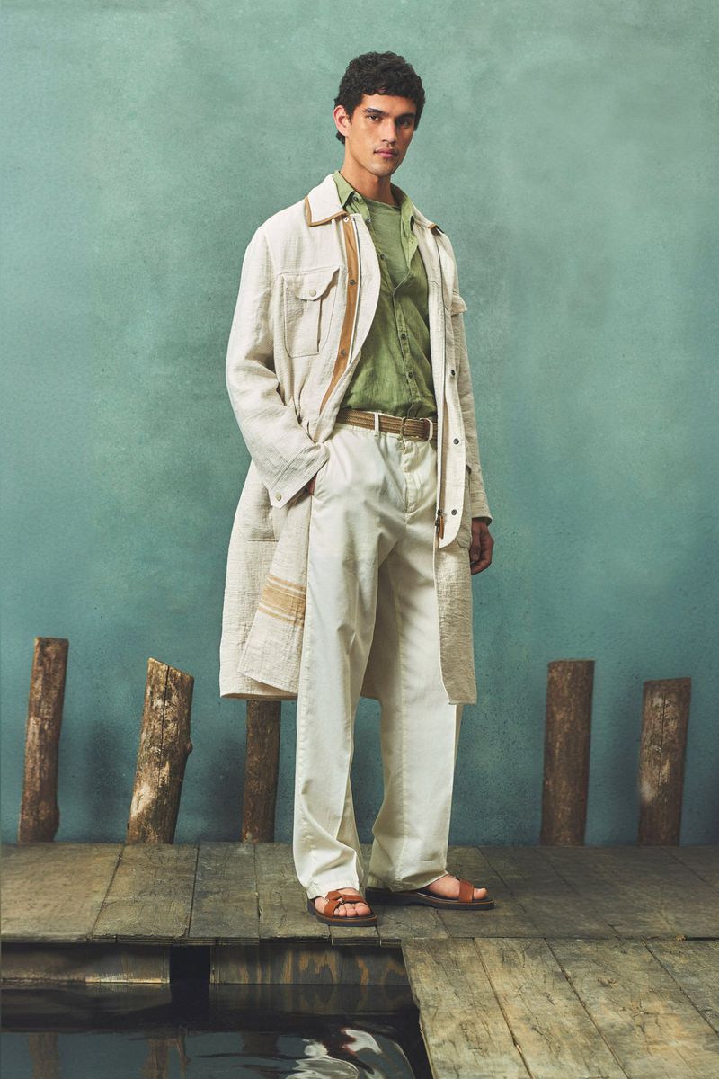 A stylish adult male poses in an unbuttoned coat and casual pants, standing on a wooden dock with a teal background.