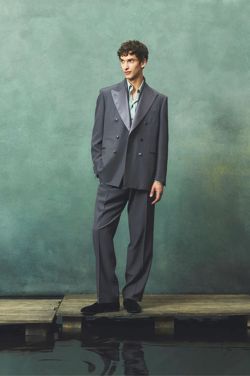 Adult male model stands on wooden planks in an elegant grey double-breasted suit and black loafers against an abstract background.