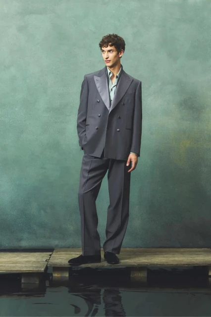 Adult male model stands on wooden planks in an elegant grey double-breasted suit and black loafers against an abstract background.