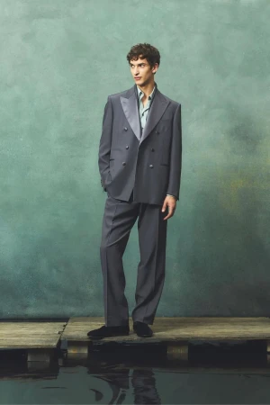 Adult male model stands on wooden planks in an elegant grey double-breasted suit and black loafers against an abstract background.