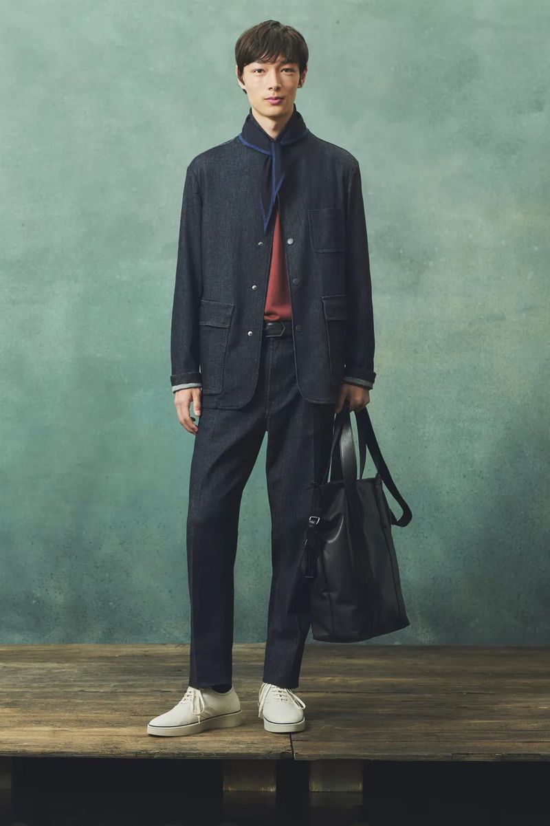 Full-length shot of a young adult male wearing a dark blue denim suit, standing on a wooden platform with a black tote bag.