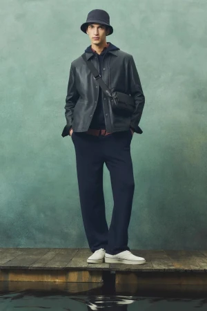 Full-length portrait of a man in a black leather jacket, blue pants, and a bucket hat standing on a wooden pier against an aqua background.