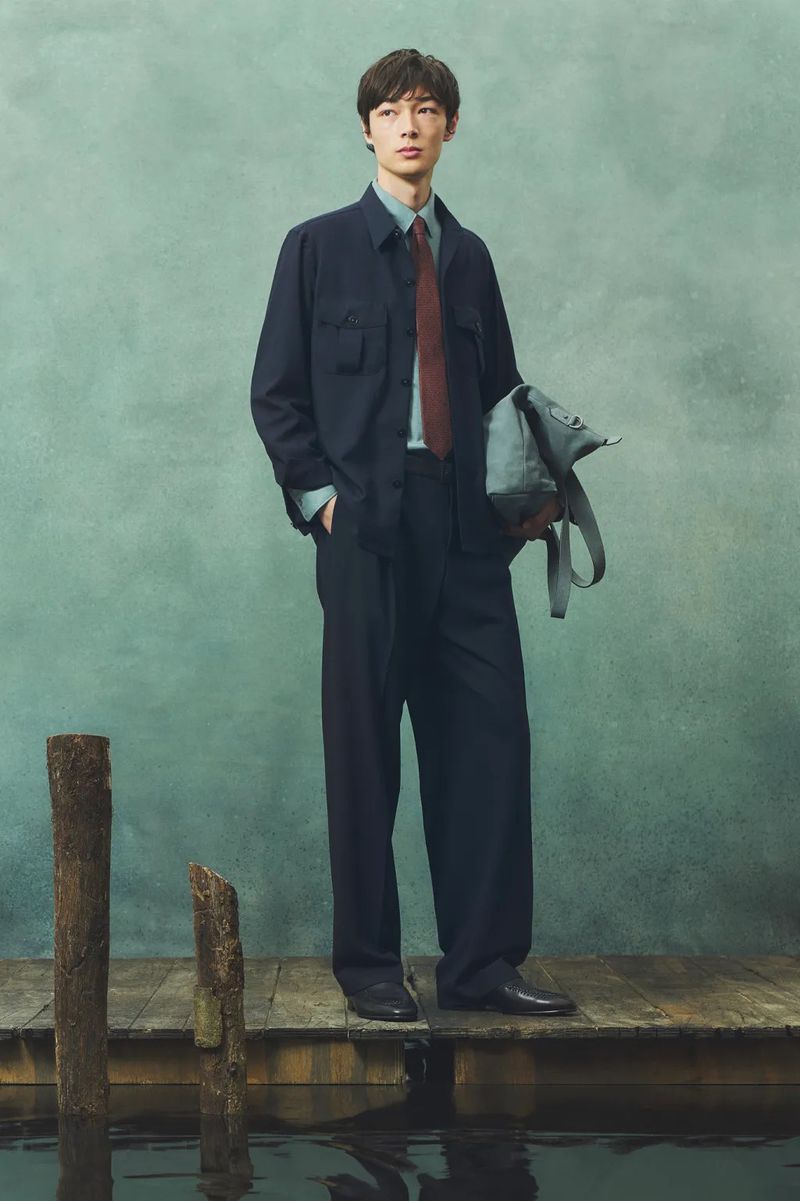 A young male model stands on a wooden pier wearing a navy suit, light blue shirt, and burgundy tie with a grey bag at dusk.