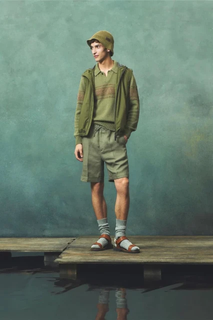 Male model in an olive green outfit, including a bucket hat, posing on a wooden dock with a blurred green background and water below.