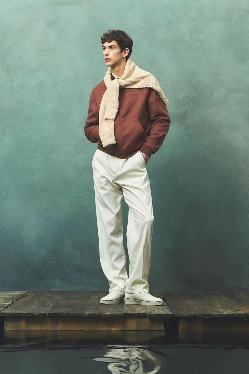 Full shot of an adult male model in rust bomber jacket with a cream scarf standing on a wooden pier on a cloudy day.