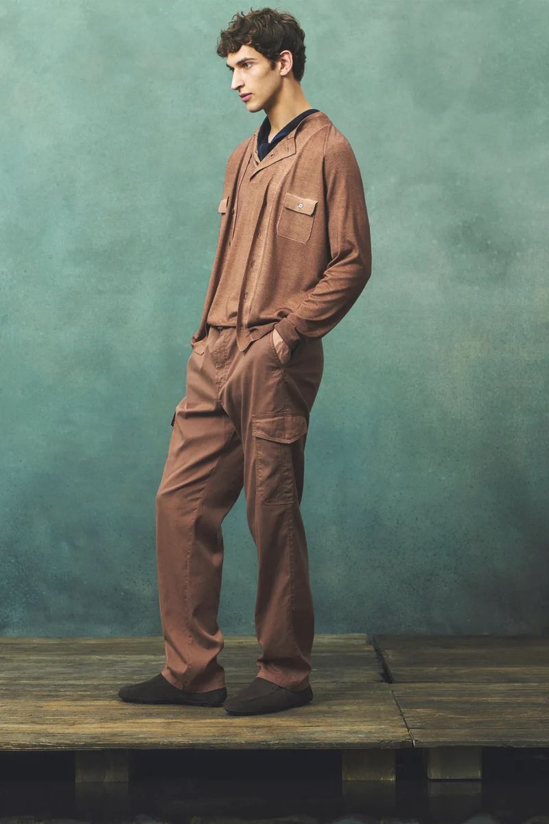 A young male model is posing in a brown jumpsuit on a wooden platform with a greenish-blue painted wall in the background, looking off to the side.