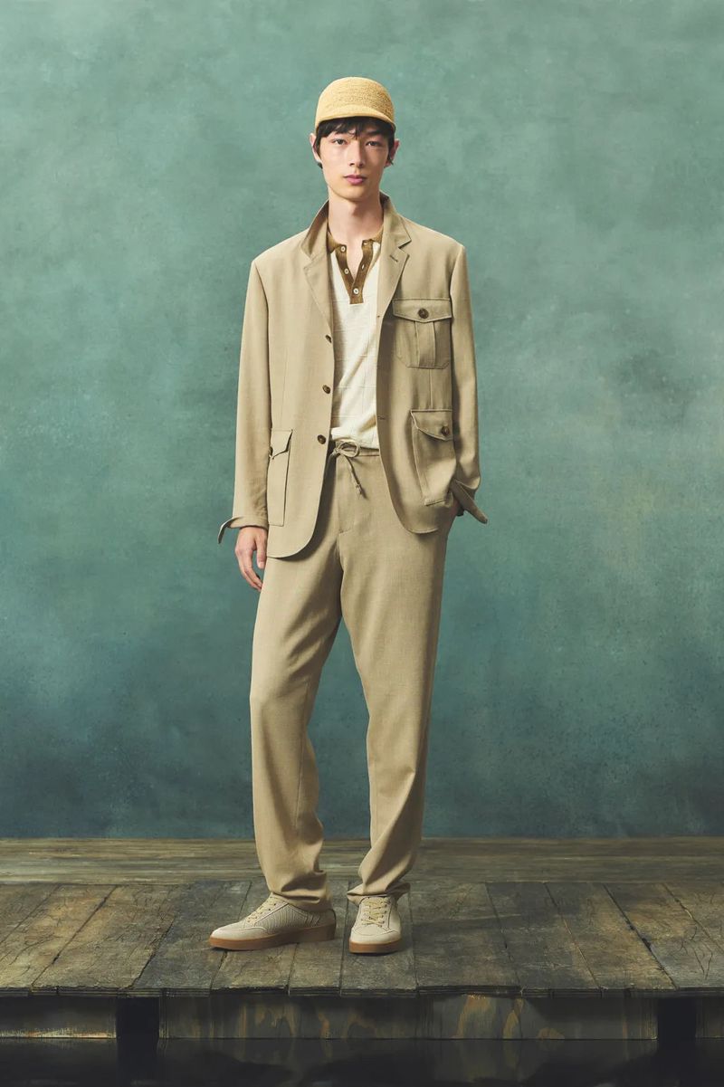 Full-length shot of an adult male model in a light brown suit and woven cap posing indoors on a wooden platform with a green backdrop.