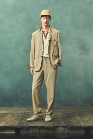 Full-length shot of an adult male model in a light brown suit and woven cap posing indoors on a wooden platform with a green backdrop.