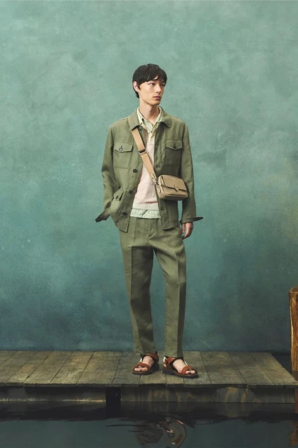 A male model poses in an olive green linen suit with a shoulder bag and brown sandals on a wooden dock with a textured blue background.