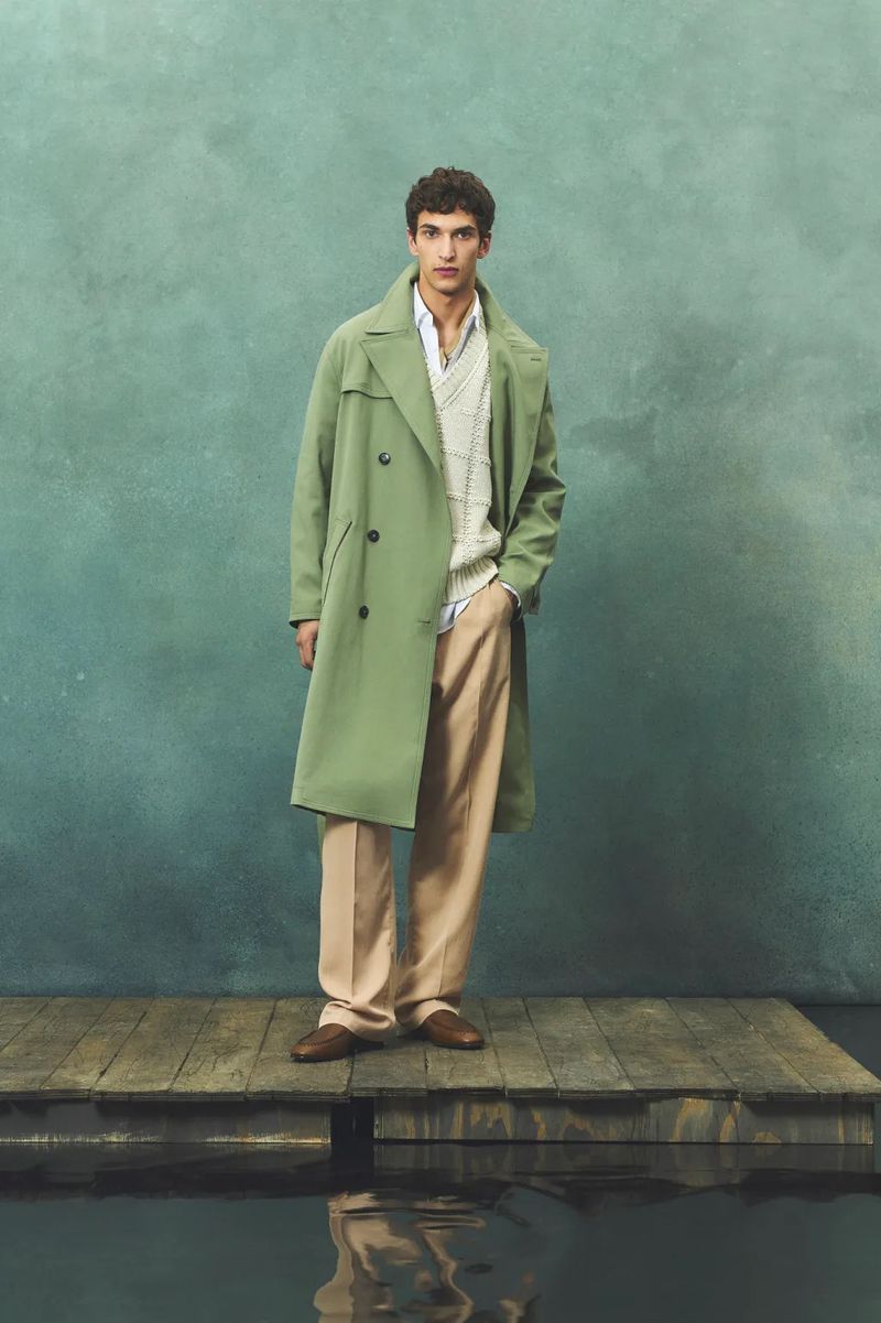 A stylish adult male stands on a wooden dock wearing a green trench coat, a patterned knit sweater, tan trousers, and brown leather shoes.