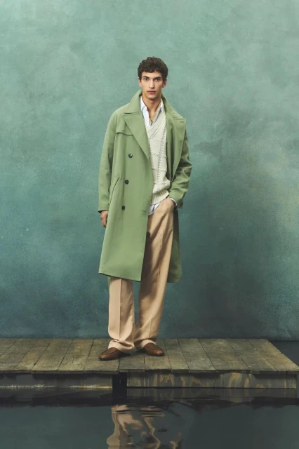 A stylish adult male stands on a wooden dock wearing a green trench coat, a patterned knit sweater, tan trousers, and brown leather shoes.