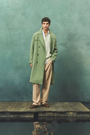A stylish adult male stands on a wooden dock wearing a green trench coat, a patterned knit sweater, tan trousers, and brown leather shoes.