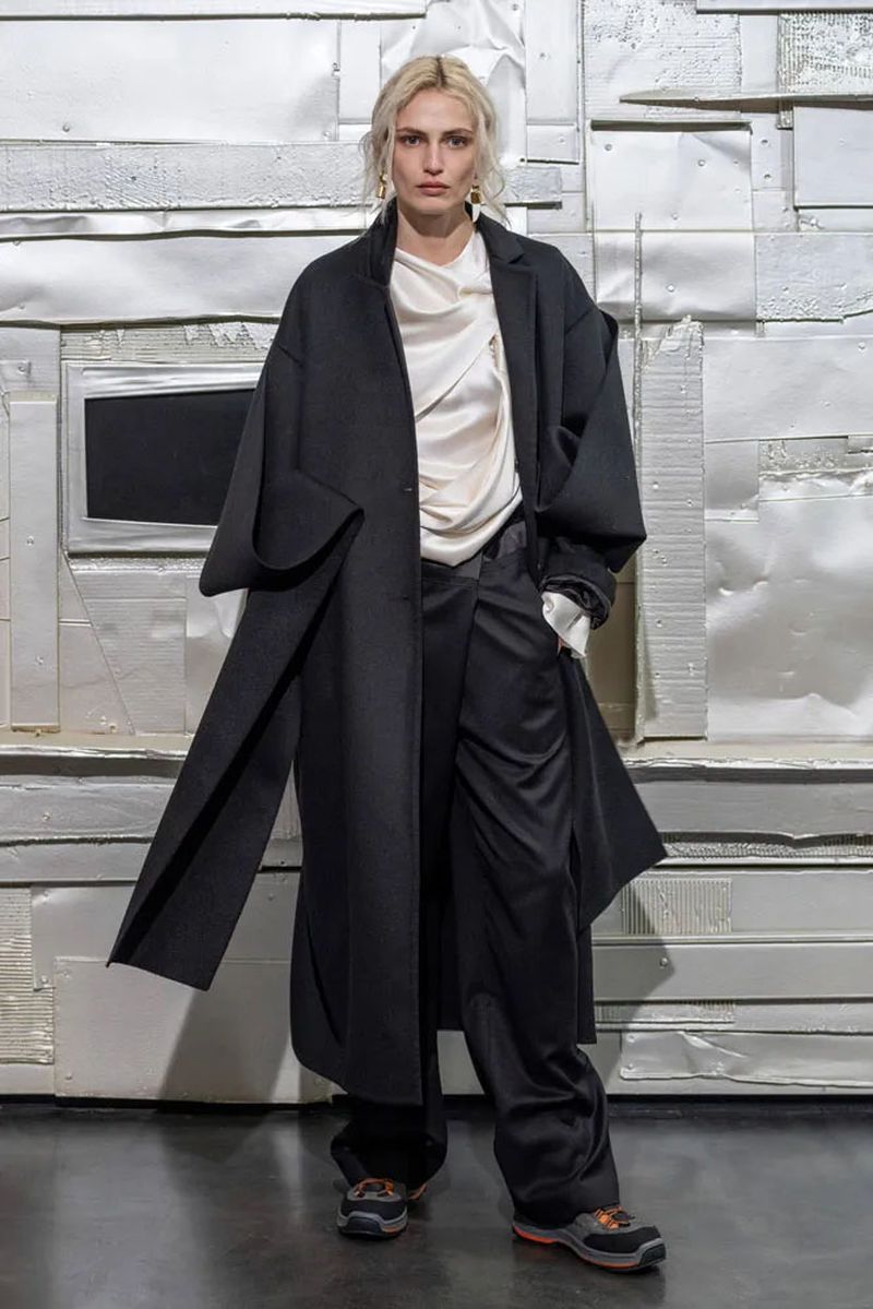 A stylish adult female stands in a black coat and pants against a textured silver background, showcasing a modern fashion aesthetic.