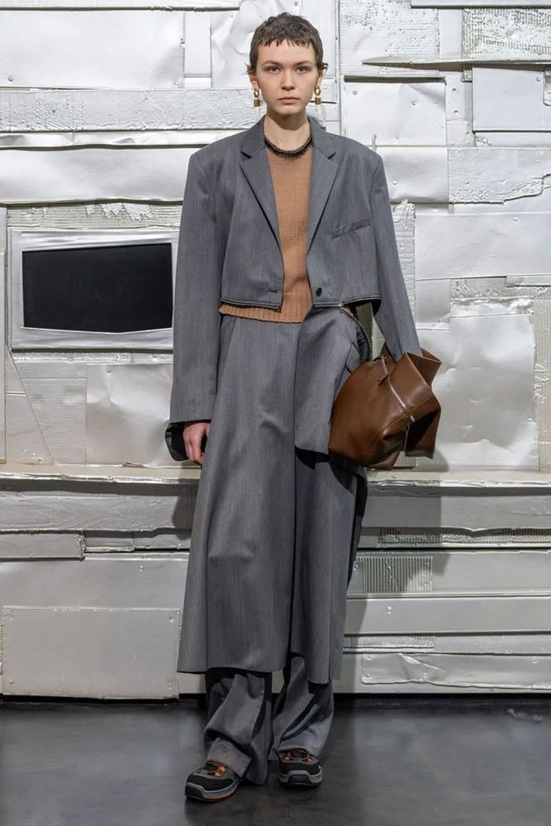 A model poses in a grey, oversized suit with a brown sweater and brown purse against a silver metal panel wall in a full-length studio shot.